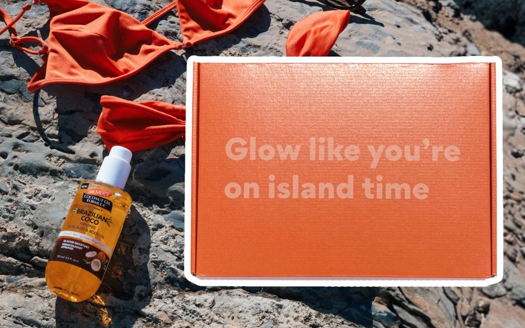 Orange bikini, sunglasses, and Brazilian Coco Firming Body Oil on rocks suggest a relaxed beach vibe. Beside them, a box reads Glow like you’re on island time.